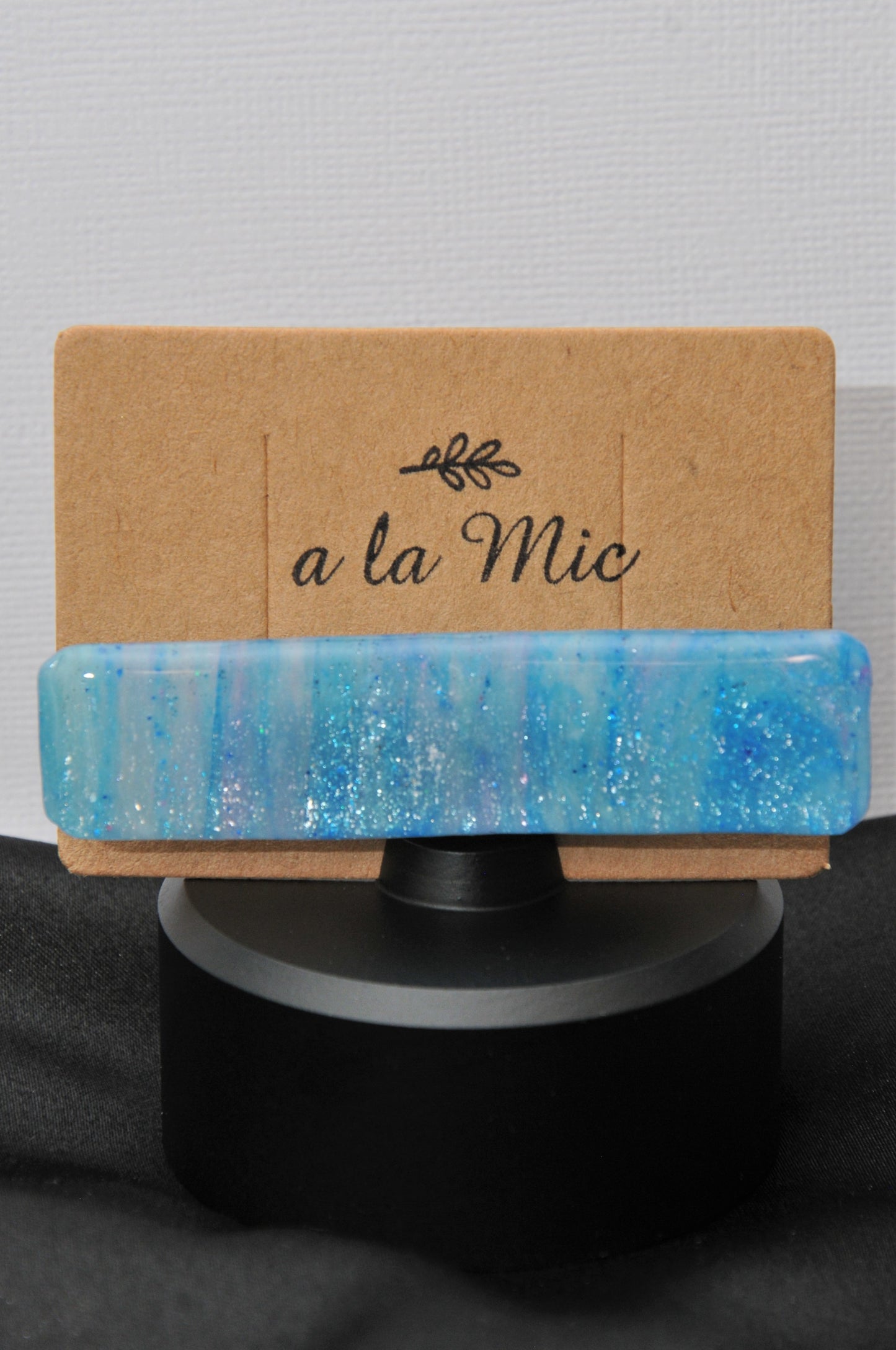 A blue and purple sparkle hair clip placed on a cardboard holder with the text 'A la mic'.